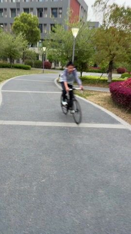 The video shows a person riding a bicycle on a paved path in a park-like setting. The person is wearing a light blue shirt and dark pants. There are trees, grass, and lampposts along the path. A building is visible in the background. The camera appears to be following the cyclist, and the view is slightly blurry due to the movement.