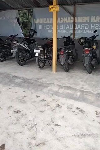 The video shows a row of parked motorcycles under a covered shelter. The shelter has a corrugated metal roof and wooden posts. A sign with text and an image of a bird is attached to a wooden post. There is a concrete ground with tire marks. Some of the motorcycles are black, one is red, and one is dark gray. The overall scene appears to be an outdoor parking area.