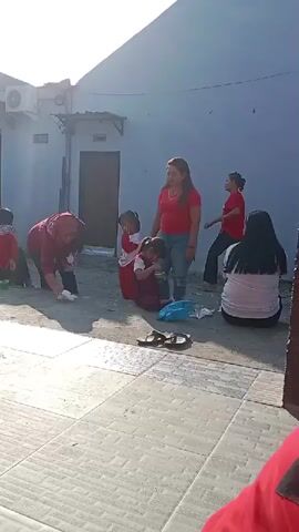 The video shows an outdoor gathering, possibly a community event or a family get-together, in what appears to be a residential area with houses in the background. The lighting suggests it is daytime, likely morning or late afternoon due to the shadows. Several people are present, including adults and children.

In the foreground, a child is sitting on a tiled floor, holding a piece of white material. Further into the scene, a woman in a red shirt and jeans stands, looking towards a child in a red and white outfit who is being held. To the left, a woman in a maroon headscarf and outfit is kneeling and appears to be interacting with something on the ground. Another person in a red shirt is also visible kneeling.

In the middle ground, there are more people seated or standing. Some are wearing red clothing. A pair of sandals is placed on the ground near some plastic bags.

The background shows the exterior of buildings, including a wall of a house, a door, and an air conditioning unit. The ground is a mix of concrete tiles and dirt or gravel.

The overall impression is of a casual, outdoor gathering with various activities taking place.