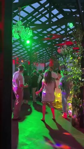 A group of people are dancing in a covered outdoor venue. Green and red lights are projected onto the floor and walls, creating a colorful and festive atmosphere. There are chandeliers hanging from the ceiling and foliage around the perimeter. The people are dressed in casual to semi-formal attire and appear to be enjoying themselves. Some individuals are dancing together, while others are dancing individually. The camera is positioned to capture the overall scene, with a focus on the dancers and the lighting effects.