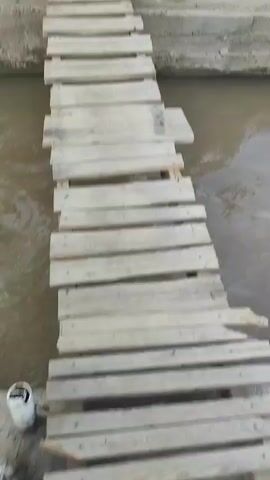 The video opens with a shaky, close-up shot of a wooden plank bridge crossing over murky water. The perspective then shifts to a wider view of a roadside scene. Two women and a child are visible, seemingly engaged in a transaction or simply resting by their goods. One woman, wearing a blue headscarf and blue jacket, holds a young boy on her lap. The boy is wearing a black t-shirt and shorts. They are seated on what appears to be an old tire. Next to them, on a low table, are an assortment of bottled drinks, including what looks like Coca-Cola and Sprite, along with bottles of cooking oil.

To the right, another woman, wearing a green hat and patterned dress, is seated and appears to be attending to her own display of bottled drinks and possibly other items. In the background, there are piles of dirt and construction materials, suggesting an area undergoing development or a construction site. Further in the distance, buildings with white walls and dark roofs are visible, along with a yellow inflatable banner that reads "Choose Healthy Milk."

The lighting suggests it is daytime, likely late morning or afternoon. The weather appears to be fair, with no signs of rain or extreme heat. The overall scene suggests a bustling, informal marketplace or roadside stall in a semi-rural or developing area, likely in Tursunzoda, Tajikistan, given the provided context. The interactions between the people seem relaxed, with the woman and child appearing comfortable together. The setting, with its mix of goods, construction, and basic infrastructure, points to a local community engaging in commerce.
