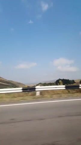 The video captures a landscape from a moving vehicle. The sky is blue with scattered white clouds. A road with a white line is visible in the foreground. Beyond the road, there is a guardrail, dry grass, and trees. Hills can be seen in the distance. A large, empty billboard structure with lights is prominent in the early part of the video, followed by a utility pole. The overall impression is of a rural or roadside environment under a clear sky.