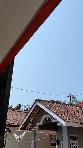 The video shows a bright blue sky with a small portion of a building's roof in the foreground. The roof is made of terracotta tiles and has white trim. There are some trees and wires visible in the background. The lighting suggests it is daytime, likely sunny.