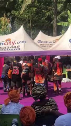 The video shows a group of people dancing and playing music outdoors on a purple-covered platform. The musicians appear to be playing traditional drums. Some of the dancers are wearing brightly colored shirts. The audience is seated and watching the performance. There are white tents with the word "Goshencity.com" printed on them. The environment appears to be a gathering or celebration. The camera is at eye-level and slightly unsteady.