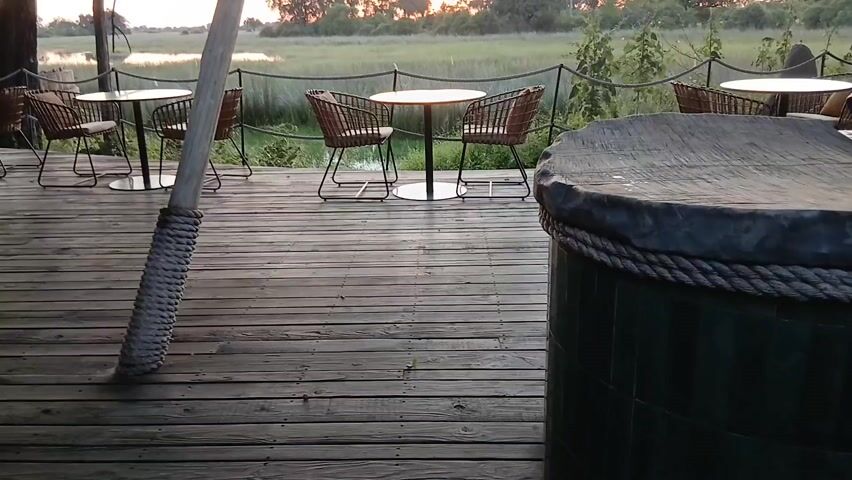 The video shows a wooden deck with several round tables and chairs arranged along the edge. The tables are small and white with silver bases, and the chairs are a light brown wicker material with dark metal frames. There is a rope railing along the edge of the deck, separating it from a grassy area that leads to a body of water. The sky is visible in the distance, with the sun setting and casting a warm glow on the landscape. In the foreground, there is a thick, rope-covered pole and a round, covered object that appears to be a hot tub or similar feature.  The scene suggests a peaceful and tranquil outdoor setting, possibly at a resort or lodge. The camera pans slightly to the right, revealing more of the deck and the surrounding landscape.