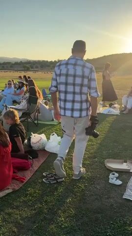 The video shows an outdoor gathering on a grassy field at sunset. The sun is low on the horizon, casting long shadows and a warm golden light across the scene. In the foreground, several groups of people are seated on blankets and chairs, some facing away from the camera. There are picnic bags, shoes, and a cardboard box on the ground. A man in a plaid shirt and light-colored pants is walking away from the camera, holding a camera in his right hand. In the background, more people are visible, some lying down or sitting. The landscape beyond the field consists of rolling hills with dry vegetation. The overall atmosphere appears relaxed and social.