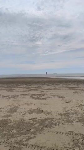 A wide shot of a sandy beach with a body of water in the background under a cloudy sky. A red lighthouse is visible in the distance on the water. There are a few small dark shapes in the water near the lighthouse, possibly birds or people. The sand in the foreground is textured with patches of darker material. The camera pans slightly to the right.