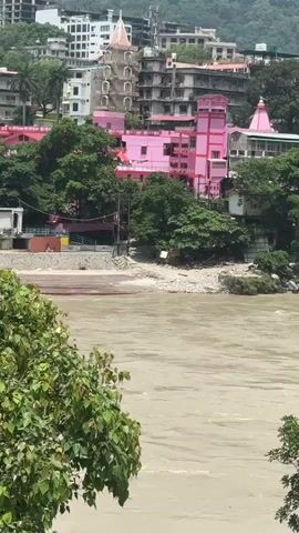 The video shows a wide, muddy river flowing in the foreground. In the background, there is a town built on a hillside, with buildings of varying heights and colors. Several trees are visible on the slopes. A prominent pink building with ornate architectural features stands out amongst the other structures, with a smaller pink structure that appears to be a temple to its right. The water in the river appears to be moving rapidly, with some debris visible along the banks. The sky is overcast, suggesting a cloudy day.