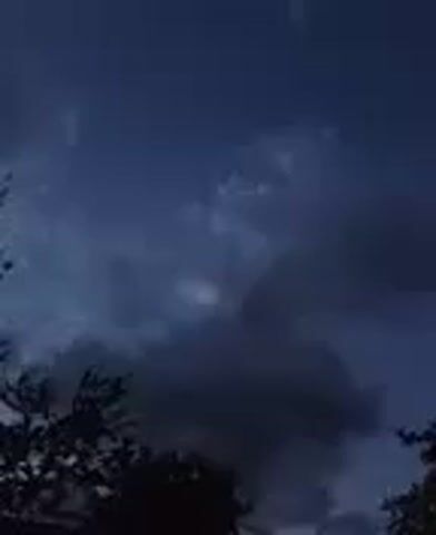 The video shows a dark, cloudy sky with silhouetted trees in the foreground. The clouds are dark and appear to be moving. The sky is a deep blue-gray color, suggesting it is either dusk or nighttime. The trees are mostly out of focus and appear to be deciduous, with some branches and leaves visible. The overall mood of the video is somber and somewhat ominous, possibly indicating an approaching storm.