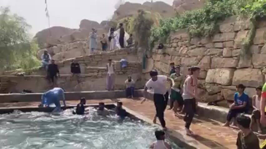 Several people are splashing around in an outdoor pool. Many are already in the water, while others stand on the pool's edge. One man in a white shirt runs and jumps into the pool, causing a large splash. There is a stone wall behind the pool with people standing and sitting on it. The scene appears to be outdoors in a natural environment, likely a hot day.