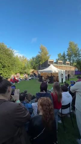 A crowd of people is gathered on a grassy lawn, facing a stage under a white tent. Some people are sitting on the grass, while others are seated on chairs. A band is performing on the stage. The sky is blue and clear, with some trees in the background. The overall atmosphere is festive and relaxed.