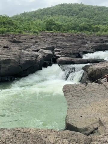 A wide shot shows a person sitting on a rocky ledge by a river. The river has strong currents and cascades over dark rocks, creating white foam. Lush green hills covered in trees form the backdrop under a cloudy sky. The person, wearing a blue shirt and dark shorts, appears to be holding a stick and looking towards the water. The rocks in the foreground are large and textured. The water is a light green color.