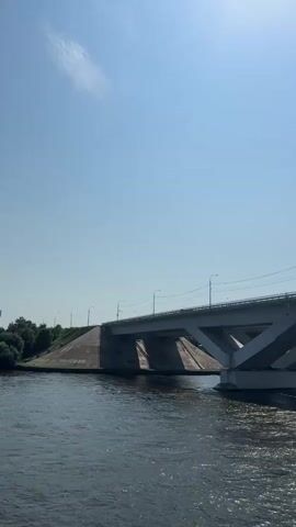 The video shows a wide river under a clear blue sky. A large, modern concrete bridge spans the river. The bridge has a distinctive triangular support structure. Cars and trucks can be seen driving across the bridge. The water in the river is dark and appears to be moving. Trees line the far bank of the river. The camera seems to be on a boat, moving towards and under the bridge.
