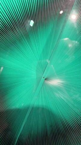 The video shows a close-up, abstract view of a textured surface illuminated by green light. The surface appears to be made of numerous thin, parallel lines that radiate outwards from a central point. There are reflections visible on the surface, suggesting it is somewhat transparent or shiny, possibly glass or plastic. The reflections include blurry shapes that could be people or objects. The lighting creates a dramatic, starburst effect due to the lines of the surface. There is a small, dark circular mark near the center of the surface. The overall impression is one of depth and movement, despite the static nature of the surface itself.
