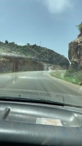 The video shows a drive through a narrow mountain pass on a paved road. The car's dashboard is visible at the bottom of the frame. The road ahead curves to the left. The rocky hillsides on either side are mostly barren with some sparse vegetation. Water is running down the cliff on the left. The sky is clear with some wispy clouds.