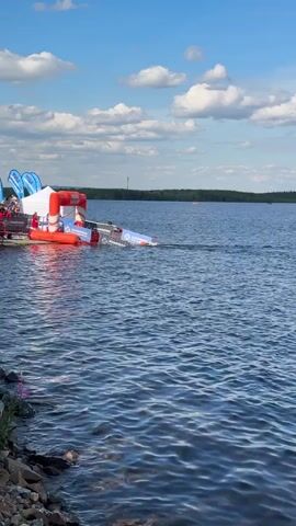 The video shows a waterfront scene on a sunny day with a blue sky and scattered clouds. In the foreground, choppy blue water laps against a rocky shoreline. In the midground, on the left side, there is an inflatable structure with an orange and white archway, and a white tent behind it. Several people are visible near the tent and the inflatable structure. To the right of the inflatable structure, a race car is partially submerged in the water and appears to be sliding down a ramp or slope into the water. The back of the car is visible, and it has orange and white markings. Further in the background, there is a body of water stretching to the horizon, with a tree line visible in the distance. There are also some buoys in the water. The overall scene suggests some kind of water race or event involving the car.