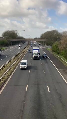 The video captures a view from an overpass looking down at a multi-lane highway, with vehicles moving in both directions. The sky is partly cloudy. Several cars, vans, and trucks are visible, navigating the highway. Trees and some greenery border the sides of the highway. Overhead streetlights illuminate the roadway. A bridge spans the highway in the distance. The camera position gives an elevated perspective of the traffic flow.