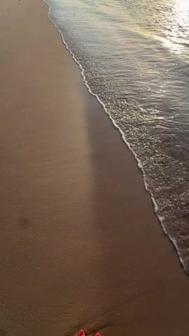 A first-person perspective video shows feet walking along a sandy beach. The camera is angled downwards, capturing the feet and the sand and water at the edge of the ocean. The water gently laps at the shore, creating a foamy white line. The sun appears to be setting or rising, casting a warm glow on the scene. The person's feet are bare, with toenails painted red. They are walking towards the right of the frame, leaving footprints in the wet sand. The environment is a beach with sand and ocean water. There are no other people or objects clearly visible in the frame. The action is walking.