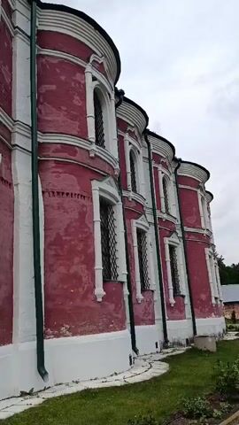 This media file features a slow pan to the right, showcasing the exterior of a grand, historical building. The building is characterized by its striking pink facade, accented with white decorative trim around its numerous arched windows. Green downspouts run vertically along the building's curved sections, adding a touch of color. The architecture suggests a religious or institutional purpose, possibly a monastery or church, given the presence of a tall, dark steeple with a cross visible in the background.

The surrounding grounds are neatly maintained, with a manicured lawn and a pathway made of interlocking bricks. Several rose bushes with vibrant pink and red blooms are visible in the foreground, adding natural beauty to the scene. Beyond the pink building, other structures with yellow facades and red-tiled roofs can be seen, hinting at a larger complex. Trees and a metal fence are also present, further defining the setting.

The weather appears overcast, with a muted sky, suggesting a cloudy day. The lighting is soft and diffused, typical of an overcast or early morning/late afternoon. No people are visible in the media. The overall impression is one of serene, historical grandeur and a well-preserved environment. The location cues, such as the architecture and the potential steeple, combined with the provided context of Radovitsy, Russia, strongly indicate a specific historical site within that region. No visible text is present in the media.