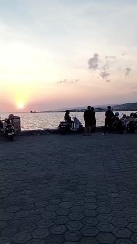 The video shows a sunset over the ocean, with a paved area in the foreground. Several people are present, some sitting on motorcycles, others standing. The sun is low on the horizon, casting a warm glow over the scene. There are a few clouds in the sky, and the water appears calm. The overall mood is peaceful and serene.