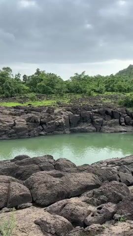 The video opens with a wide shot of a rocky riverbank under a cloudy sky. The river water is a distinct turquoise color. Large, dark rocks dominate the foreground and midground, with patches of green grass growing between them. In the distance, a dense line of green trees forms the horizon. As the video progresses, the camera pans slightly to the left, revealing more of the river and the rocky terrain. Around the 00:04 mark, a person wearing a white shirt and dark shorts is visible standing on the rocks near the riverbank, facing away from the camera and looking out towards the water and trees. The overall environment appears natural and somewhat rugged.