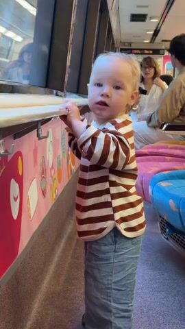 A toddler stands in a train car, holding onto a railing. The toddler is wearing a striped shirt and jeans. The train car has pink and white decorations on the wall and plush seating. In the background, two adults are visible, one holding a phone. The toddler looks toward the camera and smiles.