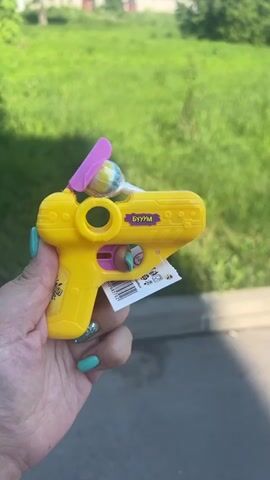 A close-up shot shows a person holding a yellow toy gun that dispenses a lollipop. The toy gun has a pink propeller on top and a colorful striped lollipop sticking out of the barrel. The background is blurred, showing green grass and a paved surface. The person's hand, with teal-colored fingernails, is visible holding the toy gun. The toy gun has some Russian text on it.