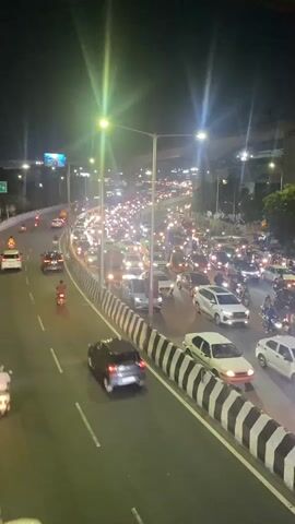The video shows a busy road at night. Many cars, motorcycles, and other vehicles are moving in the same direction on multiple lanes, creating heavy traffic. Streetlights illuminate the scene, and there is an elevated structure in the background. White and black striped barriers run along the side of the road. Some vegetation is visible in the background. The overall impression is that this is a congested urban area.