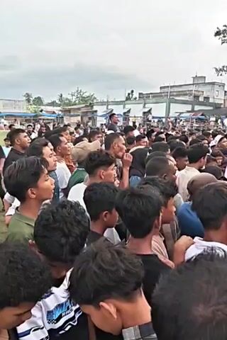 A large crowd of people, primarily men and boys, are gathered outdoors in front of a blue banner that reads "OPEN TOURNAMENT PIALA SEKJEN PAN BIREUEN" and "2023". The banner is set up on a structure that appears to be an entrance or archway. Many people are looking towards the banner and what is beyond it. Some individuals in the crowd are wearing t-shirts, while others are in more formal attire. In the background, there are tents, buildings, and trees, suggesting a public event or festival. The sky is overcast. Some people are holding mobile phones, possibly recording the event. The crowd seems to be in a state of anticipation or observation.