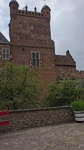 The video shows a view of a brick castle with a tall tower and ramparts. There are trees and bushes around the castle, as well as some decorative elements such as planters, statues, and a bench. The sky is cloudy. The camera pans from left to right, showing different parts of the castle and its surroundings. There are a few people visible in the distance, but they are not the focus of the video.