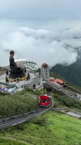 The video shows a panoramic view of a mountainous region with clouds obscuring the distant landscape. In the foreground, a red funicular train travels along a track that winds down a lush green mountainside. Above the train, on a higher plateau, is a large bronze statue of a seated Buddha, flanked by smaller statues. Adjacent to the Buddha statue is a multi-tiered structure with stone staircases and buildings, including a tall tower with a red roof. People are visible walking along the pathways and stairs of the plateau. The sky is overcast with soft clouds.