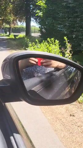 The video shows a reflection in a car's side mirror. The reflection reveals a person's hands holding a red cup or container. The person appears to be leaning out of the car window. The background in the mirror and the view from the car window show a rural or suburban environment with trees, grass, and a road. The car is in motion, as indicated by the blurred scenery in the background. The lighting suggests it is daytime.