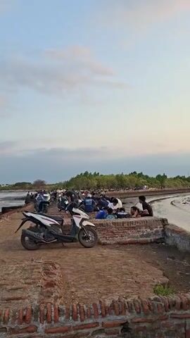The video opens on a wide shot of a muddy, shallow body of water with a treeline in the distance. In the foreground, there is a brick structure. A white and black scooter is parked on a dirt path in front of the brick structure. Several people are gathered on the path and near the water, some on motorcycles. The sky is cloudy with hints of pink and orange, suggesting it is either sunrise or sunset. The camera pans to the right, showing more people and motorcycles. The water appears to be receding, exposing mudflats. The video then moves closer to the brick structure, which shows signs of disrepair and graffiti. The camera continues to pan, focusing on the water and the distant treeline, before ending on a shot of the water and sky.