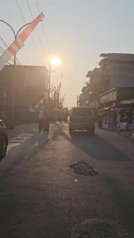 The video captures a street scene during what appears to be a late afternoon or early morning, judging by the low angle of the sun. The sun is prominently visible in the sky, casting long shadows across the asphalt road. The road itself shows signs of wear, with a noticeable pothole in the foreground. Buildings line both sides of the street, some appearing to be shops or houses. Small red and white flags, possibly national flags, are strung along the street on poles and wires, suggesting a festive or commemorative occasion. A dark-colored SUV is driving away from the camera down the street. Several motorcycles are parked along the sides of the road. In the left foreground, two individuals are walking away from the camera, one of whom is wearing a headscarf. Power lines are visible stretching across the sky. The overall atmosphere is one of everyday life in a community, possibly with a celebration in progress.