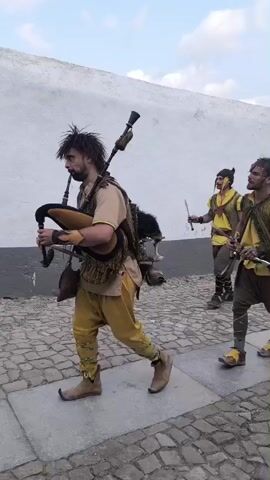 A group of people are walking down a cobblestone street. The person in the foreground is a man wearing a tan tunic, yellow pants, and brown boots. He is playing bagpipes. Behind him, another man is walking and holding two swords. Further back, a woman is spinning a hula hoop. Other people are walking behind them, some dressed in historical costumes. The buildings on the side of the street are white with some red tile roofs. There is a wall covered in purple flowers. The sky is cloudy.