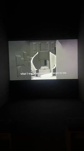 A video is displayed in a dimly lit room on a projection screen. The video shows a still, black and white architectural rendering, reminiscent of an M.C. Escher design with staircases and archways. White text appears on the screen reading, "what I was after remained obscure to me. So I would sit beside his sleeping form in one room." The projection screen is centered on the wall of the room. Two chairs are visible in the foreground. The walls of the room are bare.
