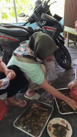 Two women are preparing food outdoors. One woman, wearing a green hijab and a light green shirt, crouches down and appears to be serving food from a plastic bag into a metal tray. Another woman, wearing a light blue hair net and a dark shirt, stands and holds a red plastic bag filled with food items and a tray of sliced cucumbers. In the foreground, there are two metal trays filled with cooked food, and a small plate with some food. Behind the women, a motorcycle is parked. The ground is paved with dark tiles. There is a wooden bench with several plastic bags on it in the background.