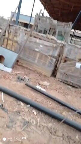The video shows a dusty outdoor scene, possibly a construction or industrial site. In the foreground, there are several long, black pipes on the ground. As the camera moves, a pile of metal scaffolding or framework comes into view. The pieces are painted with a turquoise or green color and are scattered haphazardly. Behind the metal pieces, there is a pile of what appears to be rebar, stacked in a coil. The background includes a concrete wall and a sloping hill covered with sparse vegetation. The scene is partially covered by a roof or awning. The video has the logo "itel P40 4G" in the lower left corner.