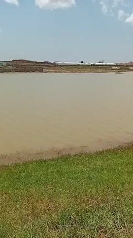 The video shows a body of muddy water with a grassy bank in the foreground. In the distance, there are several structures, possibly buildings or construction sites, and piles of dirt or sand. The sky is blue with some white clouds. The camera is stationary and pointed towards the water and the distant landscape.