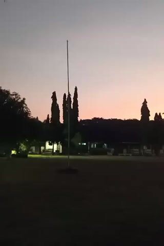 A video shows a silhouetted view of a grassy field with a flagpole in the foreground. In the background, there is a row of trees and a building with lights on. The sky is a gradient of soft pinks and oranges, indicating either sunrise or sunset. No people or specific actions are visible.