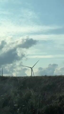 The video captures a view from a vehicle, likely a car, driving through a rural landscape under a partly cloudy sky. The foreground is dominated by tall, dry grass and some green vegetation. A prominent wind turbine stands in the middle ground, its blades slowly rotating. To the left of the wind turbine, a utility pole with wires is visible. Further in the background, more wind turbines can be seen scattered across a gentle slope. The sky transitions from a bright blue in the upper portion to darker, more dramatic clouds on the left side. The side mirror of the vehicle is visible in the bottom left corner of the frame, reflecting a glimpse of what appears to be a person's head and shoulders. The road the vehicle is traveling on is visible in the lower right of the frame, with a fence line running along the edge of a field in the distance. The overall impression is of a scenic drive through a landscape dotted with renewable energy infrastructure.