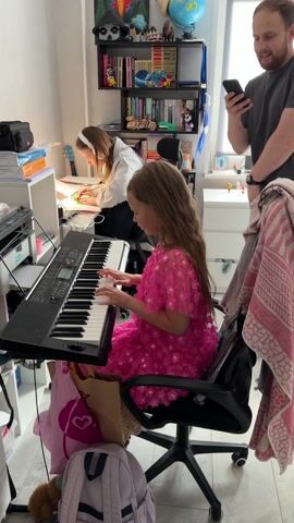 In a brightly lit indoor setting, a young girl wearing a pink dress is seated at a black electronic keyboard, playing it with both hands. Behind her on the right stands a man holding a smartphone and looking at its screen, seemingly observing the girl playing. To the girl's left, another young girl, wearing a white headband and sweatshirt, is seated at a desk, drawing or writing on a piece of paper. The room is densely furnished with a variety of objects: a tall bookshelf filled with books, toys, and decorations is visible behind the man; school supplies and bags are scattered around the keyboard; and a pink patterned towel hangs over the back of the girl's chair. The overall impression is of a cluttered but functional domestic space, with multiple activities occurring simultaneously.