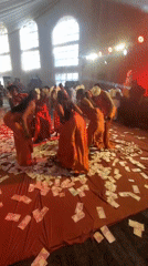 Women in orange dresses dance on money-covered stage