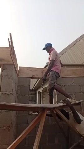 Two men are working on a construction site. One man, wearing a pink shirt, shorts, and a blue cap, is standing on a wooden scaffolding. He's holding a hammer and appears to be working on a wooden beam atop a block wall. The other man, wearing a white shirt, is positioned below on the scaffolding and reaches up to hand the man on top a brick. The building appears to be partially constructed with exposed concrete blocks. In the background, there's another building with a corrugated metal roof. The sky is overcast and bright.