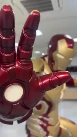 The video shows a life-sized Iron Man statue in a store. The statue is standing on a grey, rocky base, in a dynamic pose with one hand outstretched in a peace sign and the other in a defensive position. The suit is gold and maroon, with detailing that mimics the appearance of the Iron Man Mark 42 armor. The store is brightly lit with fluorescent ceiling lights and appears to be a shop with shelves full of merchandise. Several people can be seen browsing in the background. The camera zooms out to show the full figure of the Iron Man statue and its setting.