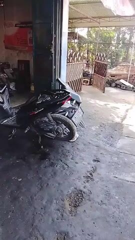 A motorcycle is parked in a cluttered workshop. The motorcycle is black and appears to be partially disassembled. The workshop floor is dirty and stained, with tools and parts scattered around. In the background, other motorcycles are visible, as well as shelves stocked with various items. The scene suggests an active repair or maintenance environment.