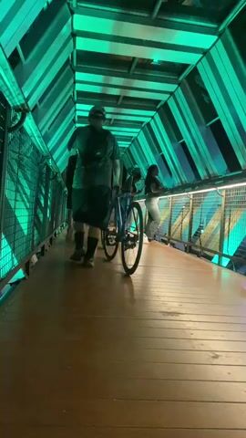 The video shows a perspective from a wooden walkway inside a tunnel-like structure. The tunnel is illuminated by changing colored lights, first green and then blue. Several people are visible on the walkway. In the foreground, a person is walking away from the camera, pushing a bicycle. Further ahead, two people are standing near the railing, one appearing to lean on it. The structure of the tunnel is visible, with exposed beams and a metal railing along the sides.