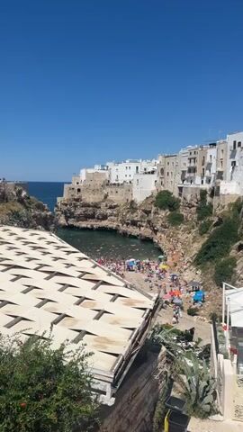 The video shows a crowded beach with many people swimming, sunbathing, and sitting under umbrellas. The beach is located in a cove with cliffs on either side. White buildings are visible on top of the cliffs. The water is clear and blue, and the sky is a bright blue. In the foreground, there is a white structure with a patterned roof.