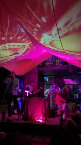 A band performs on a stage under a draped fabric illuminated with projected psychedelic visuals in pink and yellow. A male singer in a light blue jacket and black pants stands on the far left, holding a microphone. Behind him, to the right, is a keyboardist wearing a dark outfit and a face covering. A person in a colorful, patterned robe stands behind a table to the right of the keyboardist, holding a microphone. On the far right, a person with a guitar sits and plays. The stage is decorated with pink sequined fabric and flowers. The background shows a brick wall and a balcony with plants. The overall scene is dimly lit, with the projected lights creating a vibrant, atmospheric effect.