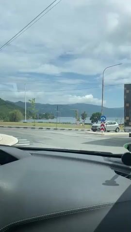 The video captures a scenic view from inside a moving car in Orșova, Romania, on a clear, partly cloudy day. The initial shot looks out the windshield over a black dashboard with a green and black phone mount. An intersection is visible with a silver SUV stopped, and a blue circular road sign is positioned nearby.

As the car turns, the perspective shifts to reveal a wide vista of the Danube River, flanked by distant green mountains under a bright, cloud-dotted sky. The view from the side window briefly shows the car's side mirror reflecting the interior, including a hand on the steering wheel.

The car then drives past a well-maintained park area adjacent to the river. This park features green grass, paved pathways, several park benches (some notably yellow), young leafy trees, and tall streetlights. A prominent white flagpole stands within the park area. The calm river stretches across the midground, with its banks lined by lush green trees, leading to the undulating mountains in the background. The overall scene depicts a tranquil and picturesque riverside landscape.